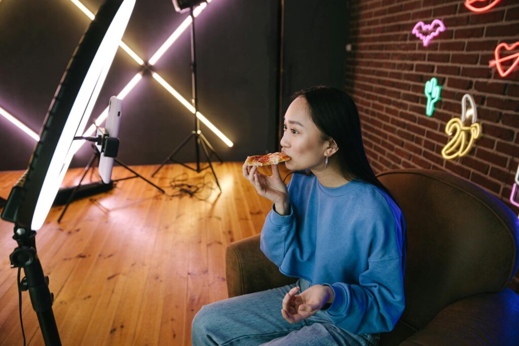 Asian woman eating pizza while streaming indoors with neon lights and ring light setup.