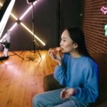Asian woman eating pizza while streaming indoors with neon lights and ring light setup.